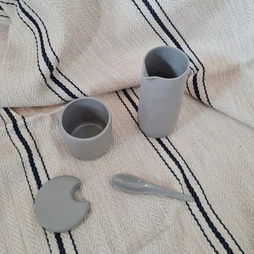 Récipient à condiments en grès avec cuillère ouvert à côté d'une cruche à eau posés sur un plaid beige
